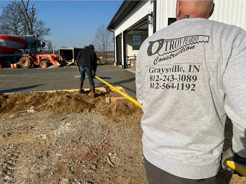 Person wearing a Troy Built Construction shirt at a construction site with a truck and open trench.