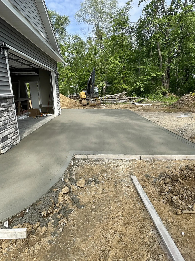 Newly poured concrete driveway in front of a house with construction equipment and trees in the background.